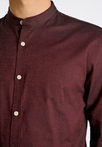 Bordeaux knoopshirt van een geweven stof met een mandarinelapel. Voorzien van vier lichtgekleurde knopen langs de sluiting.