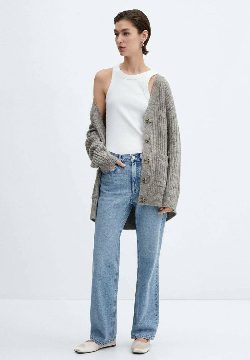 Hellgrauer gerippter Cardigan mit Knöpfen, weißes Tanktop, hellblaue gerade Jeans und cremefarbene Ballerinas, vor einem neutralen Hintergrund.
