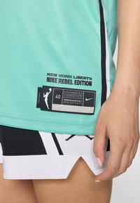 Maglia Nike New York Liberty, in tessuto mesh turchese con accenti neri, caratterizzata da un'etichetta con "Nike Rebel Edition" e un grafico di un giocatore di pallacanestro.