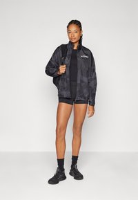 Jachetă ușoară, neagră, cu fermoar, cu un model geometric, logo din cauciuc pe piept, asortată cu pantaloni scurți sport negri și adidași.