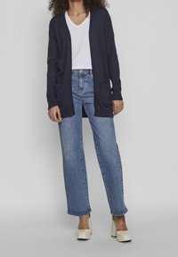 Dunkelblau gestrickte Strickjacke mit offenem Vorderteil, zwei Taschen, getragen über einem weißen V-Ausschnitt-T-Shirt und hellblauen weitem Jeans. Beigefarbene Plateau-Schuhe.