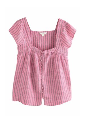 Fiatal nő göndör hajjal, rózsaszín csíkos blúzt és szürke rövidnadrágot viselve, egy strandon áll naplementekor, mögötte óceáni hullámok.