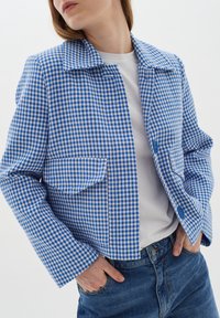 Mujer vistiendo una chaqueta a cuadros azul y blanca sobre una camisa blanca, con las manos en los bolsillos de unos jeans azules, girando la cabeza ligeramente hacia la izquierda.