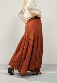 Ilgas sijonas rūdžių oranžinės spalvos, sluoksniuotas dizainas su tekstūruota medžiaga. Derinamas su juodomis sandalais. Minkštas, laisvai krentantis audinys.