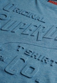 Blåt stof med hævet teksturtekst, der lyder "ORIGINAL SUPERDRY T-SHIRT CO" og en rød mærkat delvist synlig øverst.