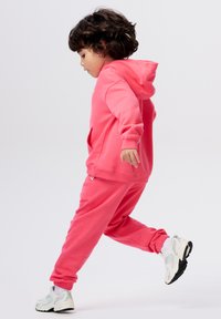 Roze hoodie met buidelzak en bijpassende sweatpants, gemaakt van zachte stof. Kind draagt witte en grijze sneakers. Zijaanzicht.