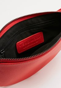 Červená syntetická taštička se zipovým uzávěrem, černým vnitřním podšíváním a viditelným značkovým štítkem. Hladký povrch s zaobleným tvarem.