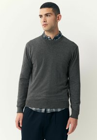 Suéter gris de punto con cuello redondo, puños y dobladillo acanalados. Llevado sobre una camisa de cuadros azul y blanca, con pantalones oscuros visibles.