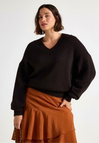 Žena s krátkými hnědými vlasy má na sobě černý svetr s véčkovým výstřihem a oranžovou sukní s volánky a drží černý řetízek se zlatými detaily.