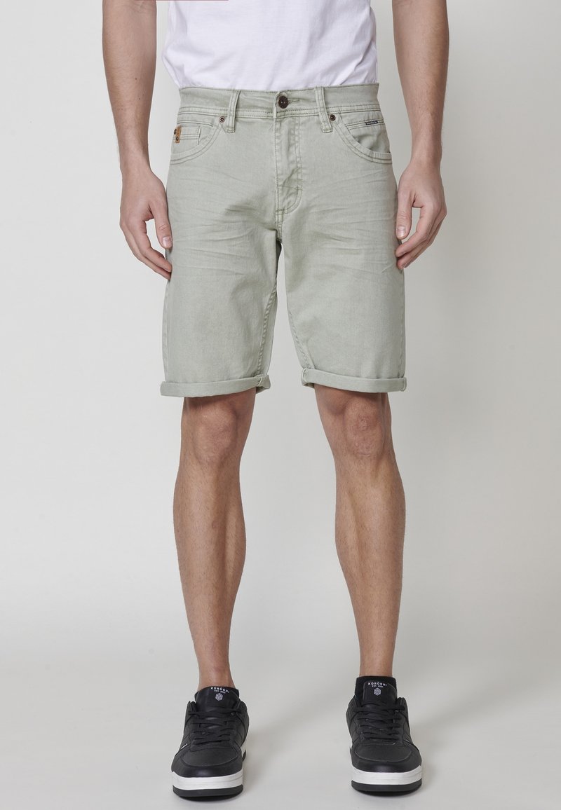 Moški v svetlo zelenih zavihanih skinny hlačah, črnih supergah in belem majici, ki stoji ob enostavni svetli ozadju.