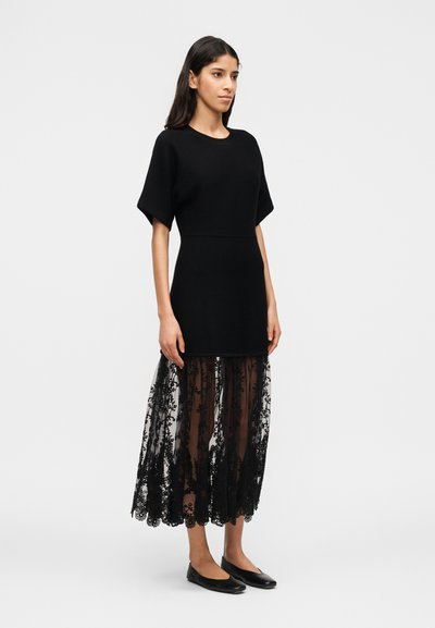 Γυναίκα στέκεται φορώντας μαύρο φόρεμα με κοντά μανίκια, διάφανο κεντητό δαντελένιο φούστα και μαύρα φλατ παπούτσια μπροστά σε απλό φωτεινό φόντο.