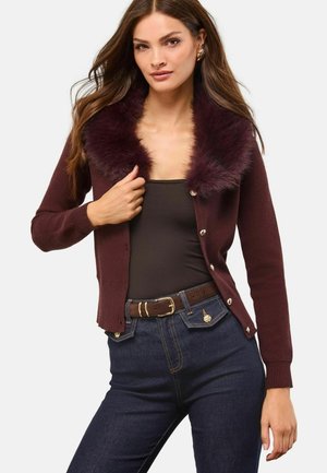 Burgunderfarbener Cardigan mit einem Kunstfellkragen, ausgestattet mit Knopfverschlüssen, gerippten Bündchen und einem taillierten Schnitt, getragen über einem schwarzen Top und Jeans.