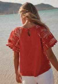 Blusa rossa con maniche ricamate, caratterizzata da un dettaglio con bottone a chiave sul retro, vestibilità morbida e una texture soffice.