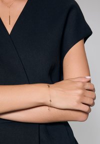 Złota bransoletka z małymi ozdobnymi charmsami, delikatnym łańcuszkiem, noszona na lekko opalonej nadgarstku, dopełniająca czarny strój.