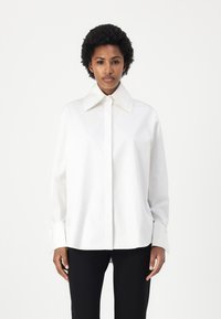 White button-up shirt with a large collar, long sleeves, and a relaxed fit. Made of smooth fabric, features a subtle sheen and cuff detail.