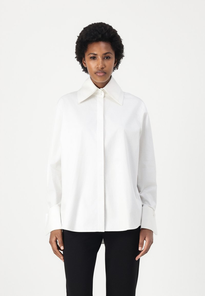 White button-up shirt with a large collar, long sleeves, and a relaxed fit. Made of smooth fabric, features a subtle sheen and cuff detail.