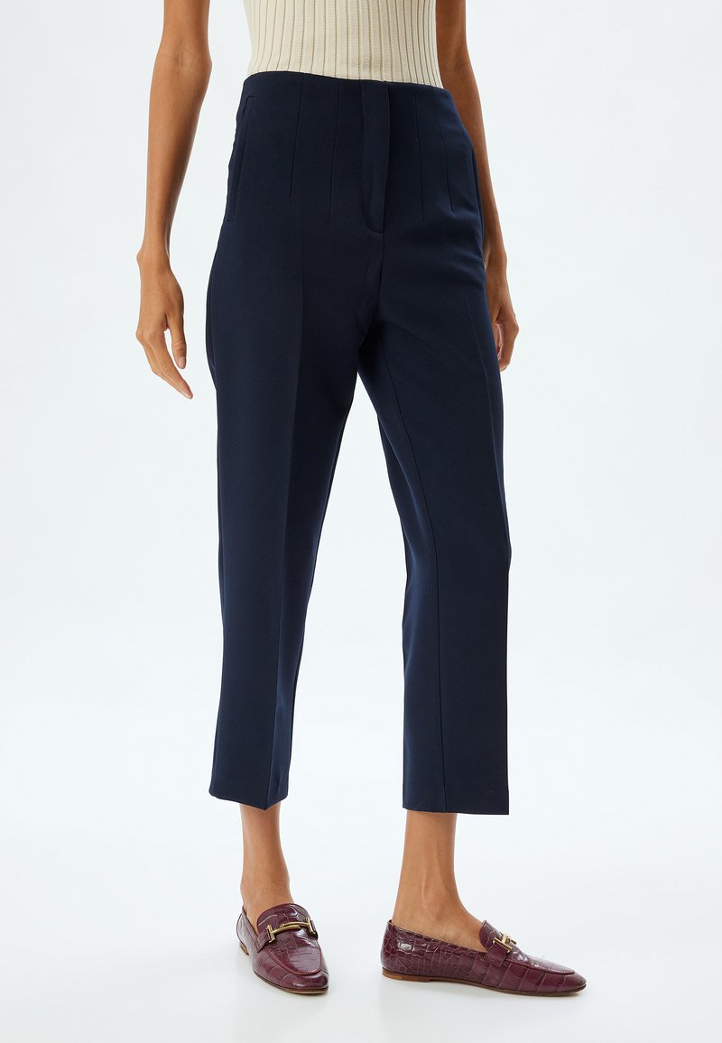 Pantalons taillés bleu marine avec plis avant et poches latérales, présentant une longueur courte. Portés avec des mocassins décoratifs bordeaux.
