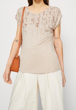 Vrouw draagt een beige top met korte mouwen en patroon, witte broek met fijne strepen en een bruine schoudertas, toont tatoeages op beide armen.