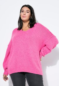 Jasnoróżowy, oversize sweter z dekoltem w serek, o miękkiej fakturze, z prążkowanymi mankietami i dołem. Noszony z ciemnymi dżinsami na białym tle.