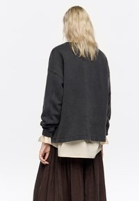 Sudadera gris oscuro con hombros caídos, costuras mínimas y puños de canalé, superpuesta sobre una camisa color crema y una falda marrón.