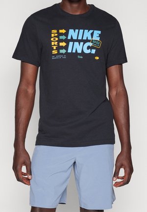 Mężczyzna ubrany w czarną koszulkę z żółto-niebieskim napisem "SPORTS NIKE INC." i jasnoniebieskie spodnie, stojący na jednolitym tle.