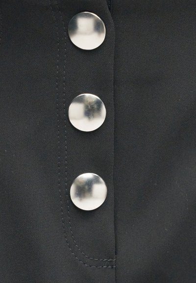 Τρία γυαλιστερά ασημένια στρογγυλά κουμπιά σε σκούρο ύφασμα με ορατή ραφή κατά μήκος του κουμπωτού ανοίγματος.