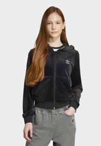Juodas velūrinis zip-up džemperis su priekiniais kišenėmis, gumbeliais ant rankovių ir mažuoju baltu logotipu kairėje krūtinės pusėje, derinamas su pilkomis elastinėmis kelnėmis.