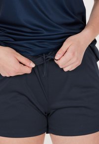 Person som drar åt snöret på mörkblå sportshorts, iförd en matchande blå topp, mot en enfärgad bakgrund.