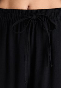 Un pantalon noir à cordon de serrage présente une taille froncée avec un détail de nœud. Le tissu semble doux avec une texture lisse et une coupe décontractée.