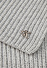 Un tessuto a costine di un grigio chiaro presenta un piccolo accento con logo metallico. La texture è morbida, con evidenti coste verticali.