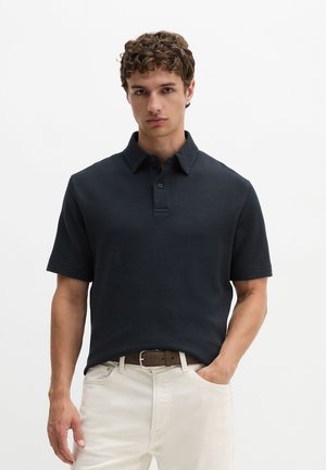Jonge man met krullend haar die een donker gestructureerd polo met korte mouwen en lichtbeige broek met een donkere riem draagt, staand met één hand in de zak.