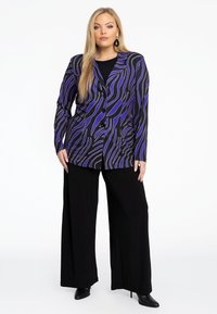 Blazer multicolore avec un motif ondulé violet et noir, à un bouton, associé à un pantalon large noir taille haute.