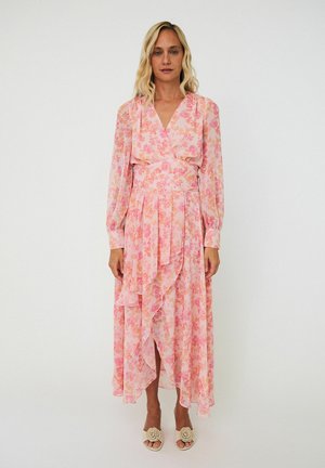 Frau trägt ein langärmeliges, rosa geblümtes Wickelkleid mit Rüschen-Details und beige Sandalen mit Blumenakzenten, steht vor weißem Hintergrund.