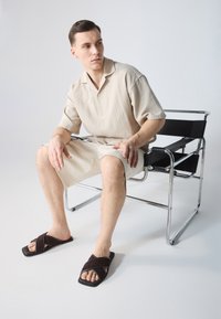 Beige kortermet skjorte, lyse kremshorts og mørkebrune tversover sandaler. Modell sitter på en stol med kromramme. Glatte teksturer.