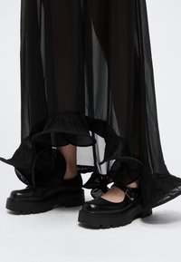 Sorte chunky Mary Jane-sko båret med en gennemsigtig sort nederdel med flæsekant, vist på lysegrå baggrund.