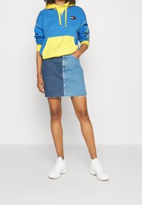 Sudadera bloqueada en azul y amarillo con un bolsillo, combinada con una falda de mezclilla de tonos bicolor y dobladillo deshilachado. Zapatillas altas blancas.