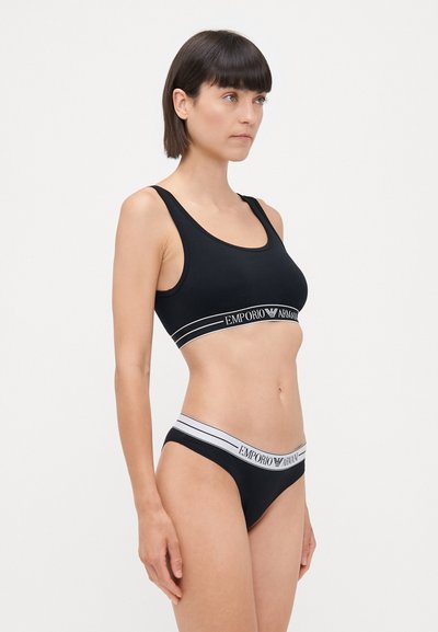Black cotton sports bra with a wide elastic band featuring white stripes and logo. Matching black thong with similar elastic band design.