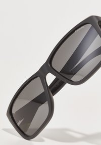 Ochelari de soare din plastic negru cu rame mari pătrate și lentile întunecate. Brandul Polaroid este inscripționat în colțul din stânga sus al lentilei drepte.
