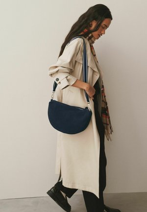 Kobieta w beżowym trenczu i kraciastym szaliku niesie granatową zamszową torbę na ramię, idąc wewnątrz budynku na tle jednolitej ściany.