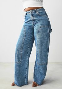 Jeans a gamba larga in denim chiaro con tasche a toppa, orli sfilacciati e un lavaggio sbiadito. Presentano un vita alta e una chiusura frontale con bottone.