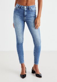 Ψηλόμεσα στενά τζιν σε ανοιχτό μπλε ντένιμ, με λεία υφή, πέντε τσέπες και αντίθετες ραφές.