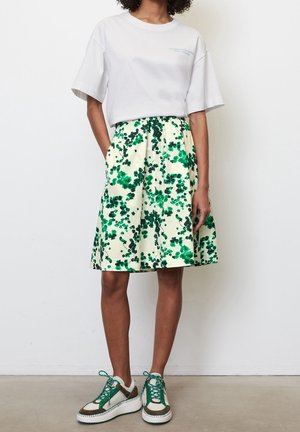 Witte cropped T-shirt met bedrukte tekst. Bloemenknielange rok in crème met groene patronen. Groene en bruine sneakers met witte zolen.