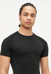 Schwarzes Sportshirt mit einem strukturierten dreieckigen Muster auf der Brust, körpernahem Schnitt und kurzen Ärmeln. Glattes, dehnbares Material.