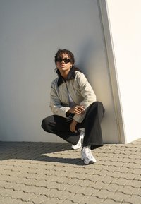Giacca trapuntata beige con colletto nero, t-shirt grigia, pantaloni neri a gamba larga e sneakers bianche; persona con capelli ricci e occhiali da sole.