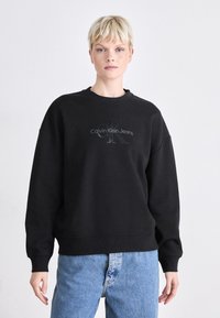 Svart genser med rund hals og senket skuldre, med en tonet Calvin Klein Jeans-logo foran. Brukt med blå jeans.
