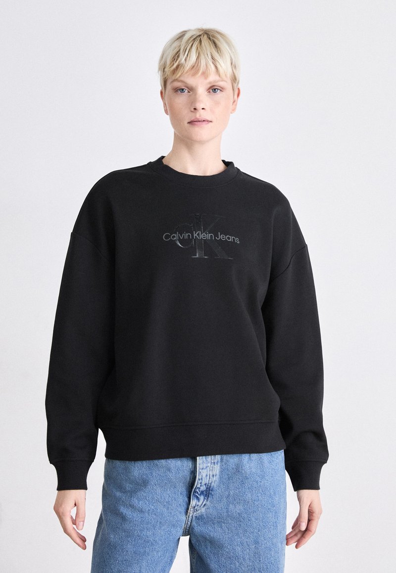 Svart genser med rund hals og senket skuldre, med en tonet Calvin Klein Jeans-logo foran. Brukt med blå jeans.