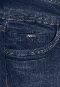 Mörkblå denimjeans med synlig sömnad i orange. Har en bakficka med en liten "Pepe Jeans"-etikett fäst.