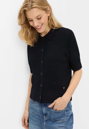 Blond kvinde smiler, iført sort kortærmet knap-up striktrøje og blå denim jeans, hånd i lommen mod hvid baggrund.