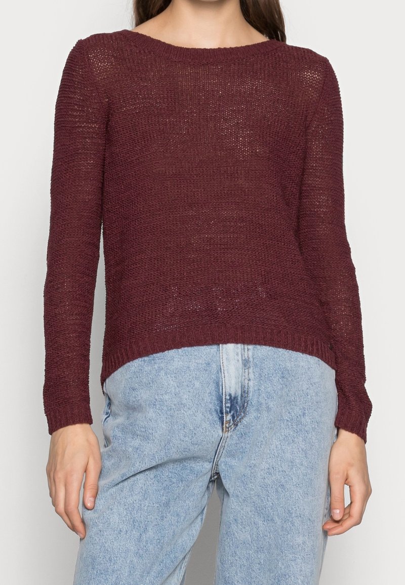 Femme portant un pull en tricot bordeaux à manches longues et un jean taille haute bleu clair sur un fond uni.