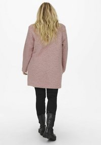 Manteau texturé rose à manches longues, tombant jusqu'à mi-thigh, associé à un pantalon noir ajusté et des bottes noires mi-mollet. Aucune fermeture visible.
